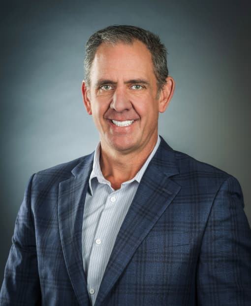 Professional headshot of Derek Panfil, wearing a navy blue plaid blazer and striped dress shirt, smiling against a gray studio background