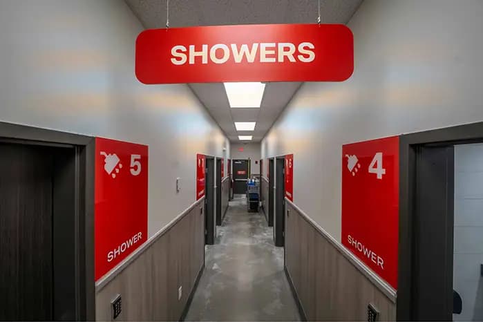 A hallway of numbered individual shower stalls (visible stalls 4 and 5 in foreground) with a large red overhead sign reading 'SHOWERS' and additional stalls continuing down the corridor