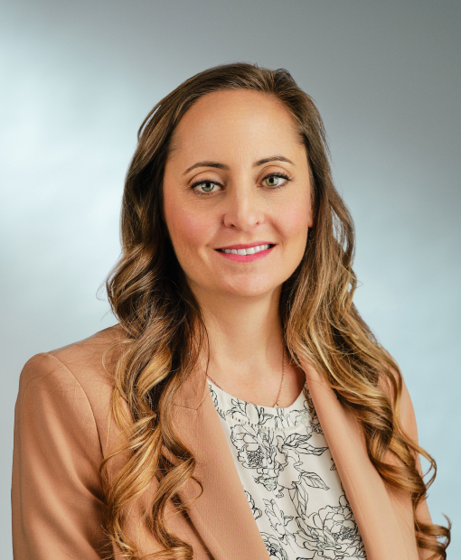 Professional headshot of Adrienne Ingoldt, a woman with long wavy brown highlighted hair, wearing a peach blazer over a white floral blouse, smiling against a light gray background