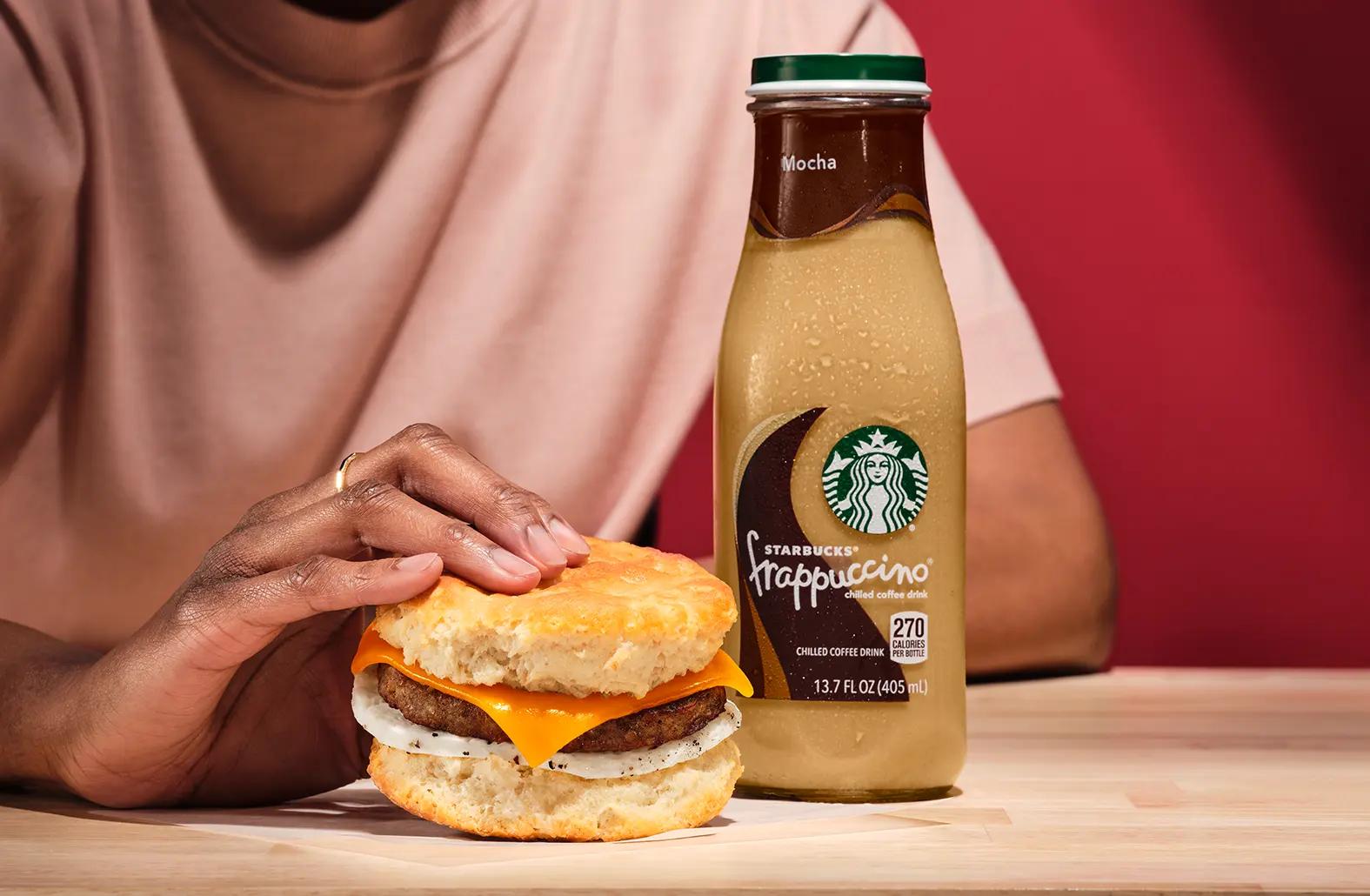 Person enjoying a sausage, egg and cheese biscuit and a Starbucks Frappuccino.