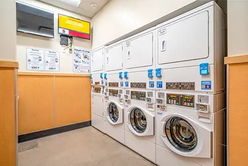 A commercial laundry room with a row of front-loading washing machines topped by stacked dryers, each unit equipped with card payment and control panels, with instructional signage and a payment kiosk mounted on the wall