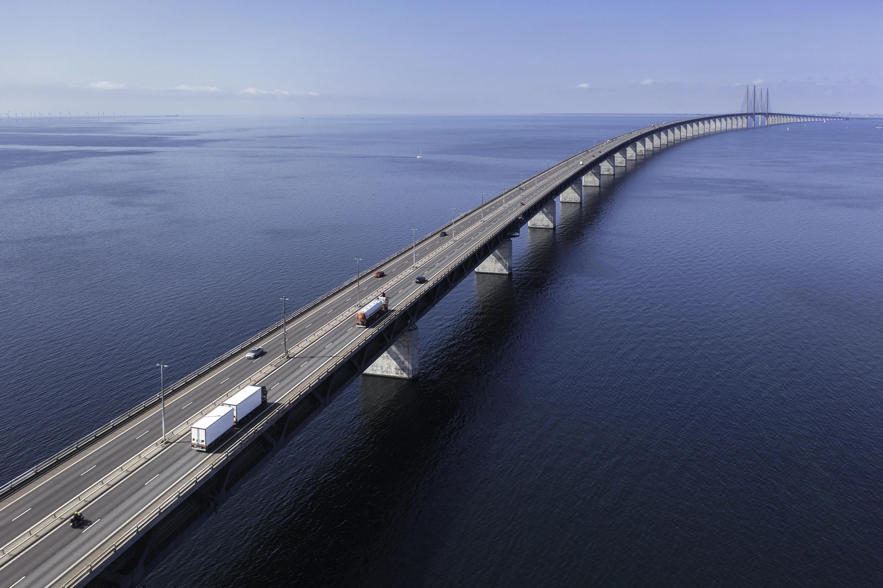 Aerial view of a long multi-lane bridge stretching across a calm blue sea toward the horizon, with trucks and cars in transit and cable-stay towers visible in the far distance