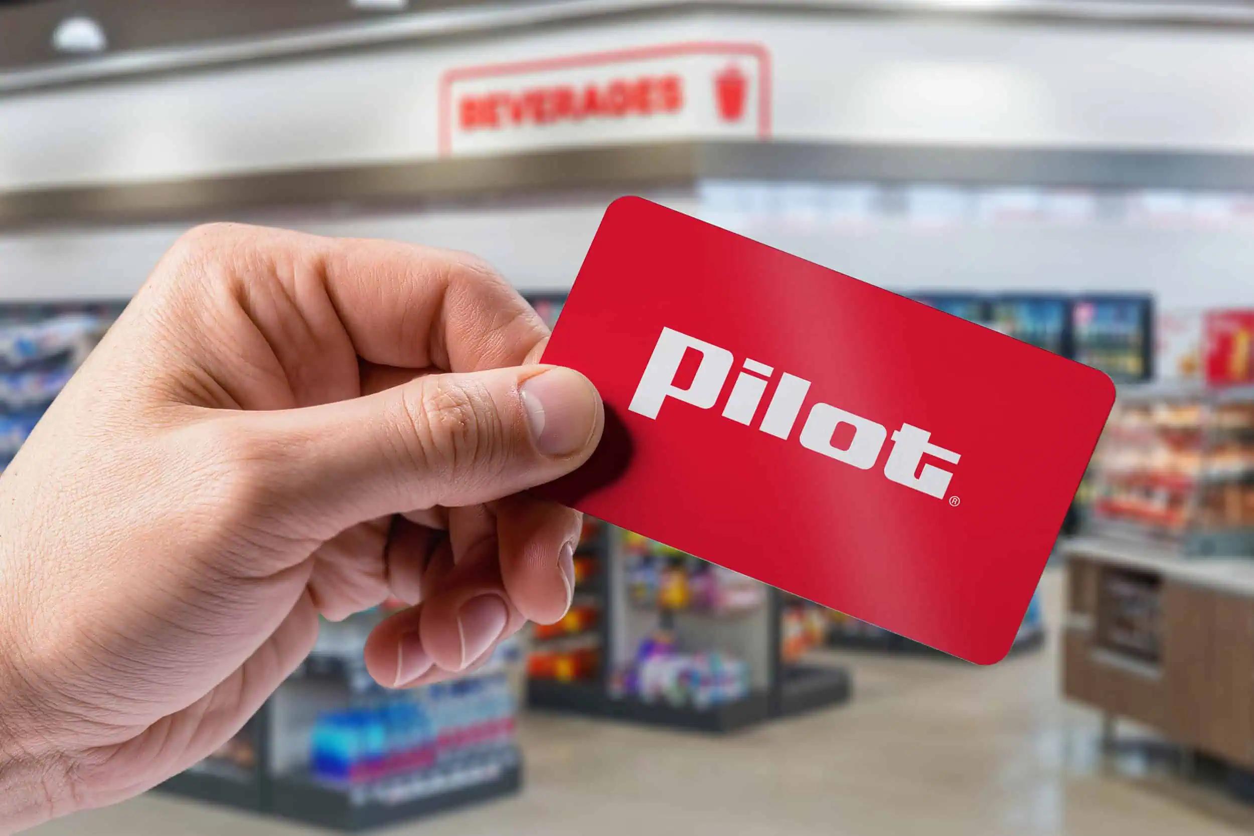 Hand holding a Pilot gift card with the Pilot logo on a red background inside a Pilot travel center convenience store.