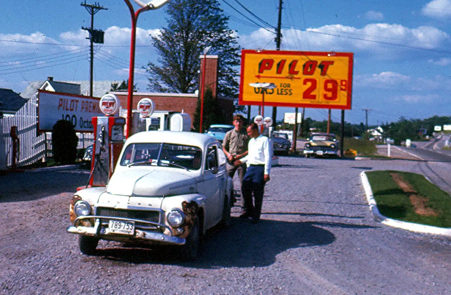 Vintage 1960s photo of a Pilot gas station with gas priced at 29.9 cents per gallon; two men stand beside a white Volvo PV544 sedan at the gravel station forecourt, with gas pumps and a large yellow Pilot 'Gas For Less' sign visible in the background