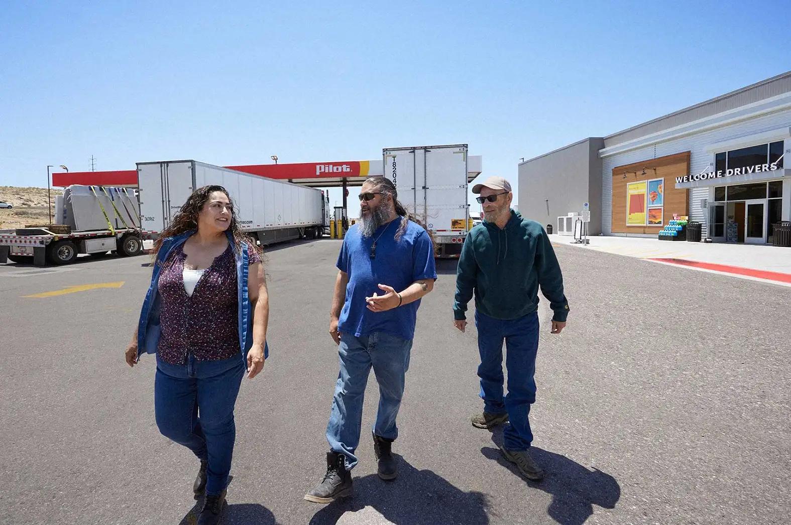 Three truck drivers walking across the parking lot of a Pilot Flying J travel center, with semi-truck trailers and a 'Welcome Drivers' sign visible in the background