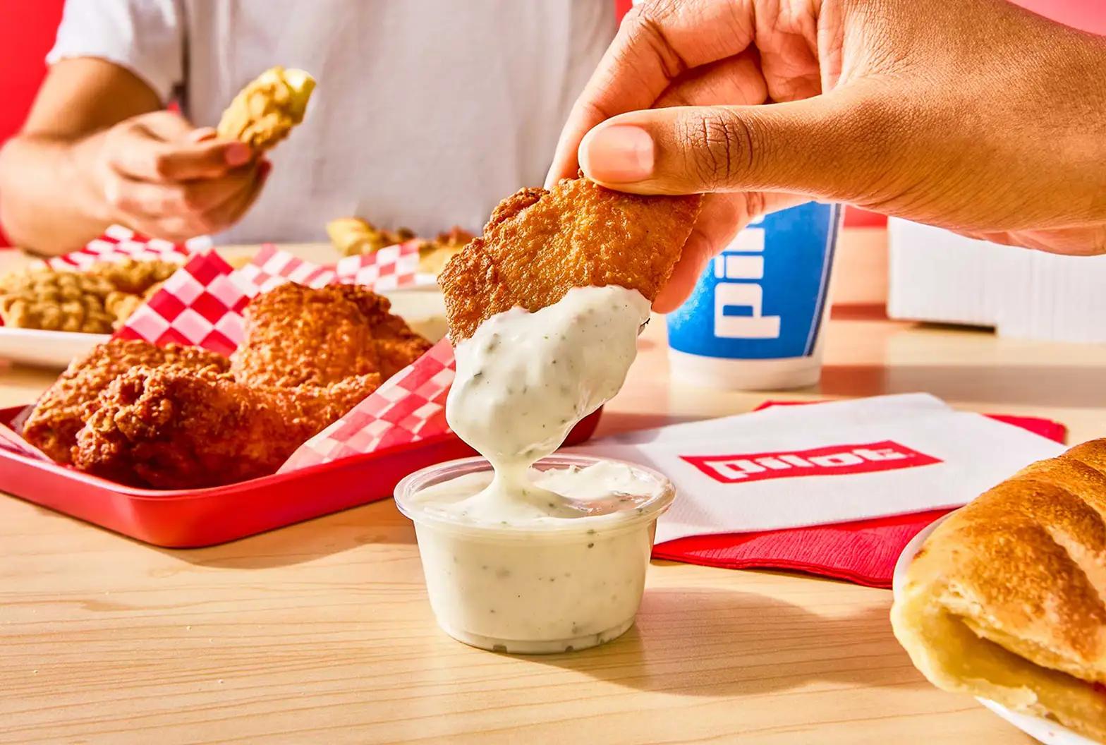A hand dipping a crispy fried chicken wing into a cup of ranch dressing, with a red tray of fried chicken pieces in checkered paper, a biscuit, and a Pilot branded drink cup on a wooden table