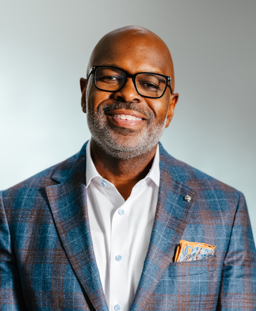 Julius Cox, a smiling man wearing black-framed glasses, a blue plaid blazer, white dress shirt, and an orange paisley pocket square
