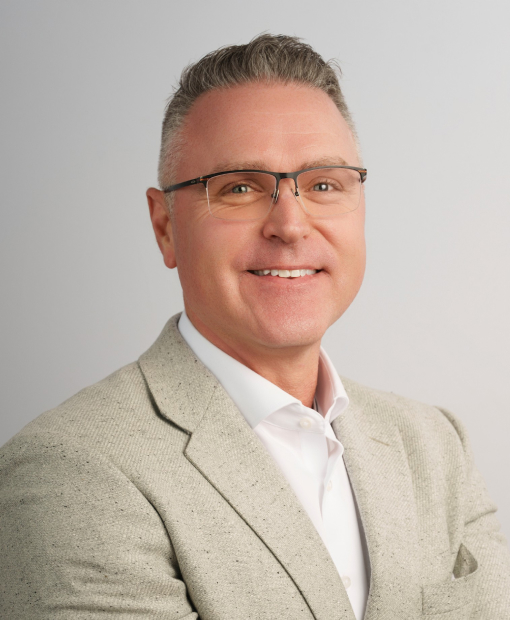 Professional headshot of Shannon Sturgil, smiling man wearing glasses and a light beige blazer over a white dress shirt