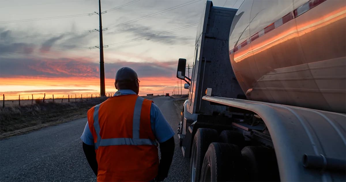 A worker in an orange high-visibility safety vest walks alongside a large tanker truck on a rural road at sunrise, with a colorful orange and red sky and power lines visible in the background
