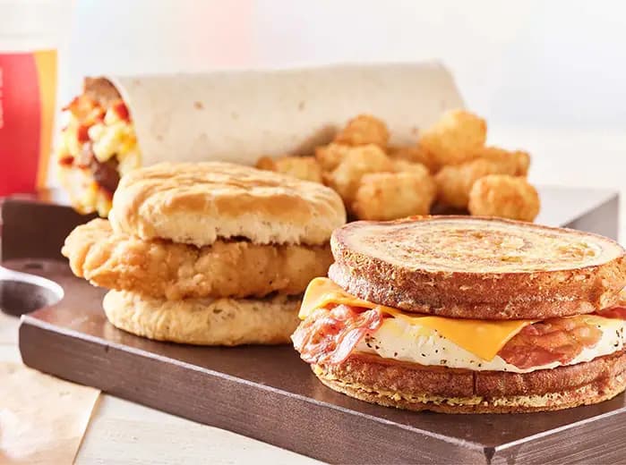 A breakfast spread on a wooden tray featuring a toasted multigrain sandwich with bacon, egg, and cheese in the foreground, a fried chicken biscuit sandwich, a breakfast burrito, and a side of tater tots, with a drink cup in the background.