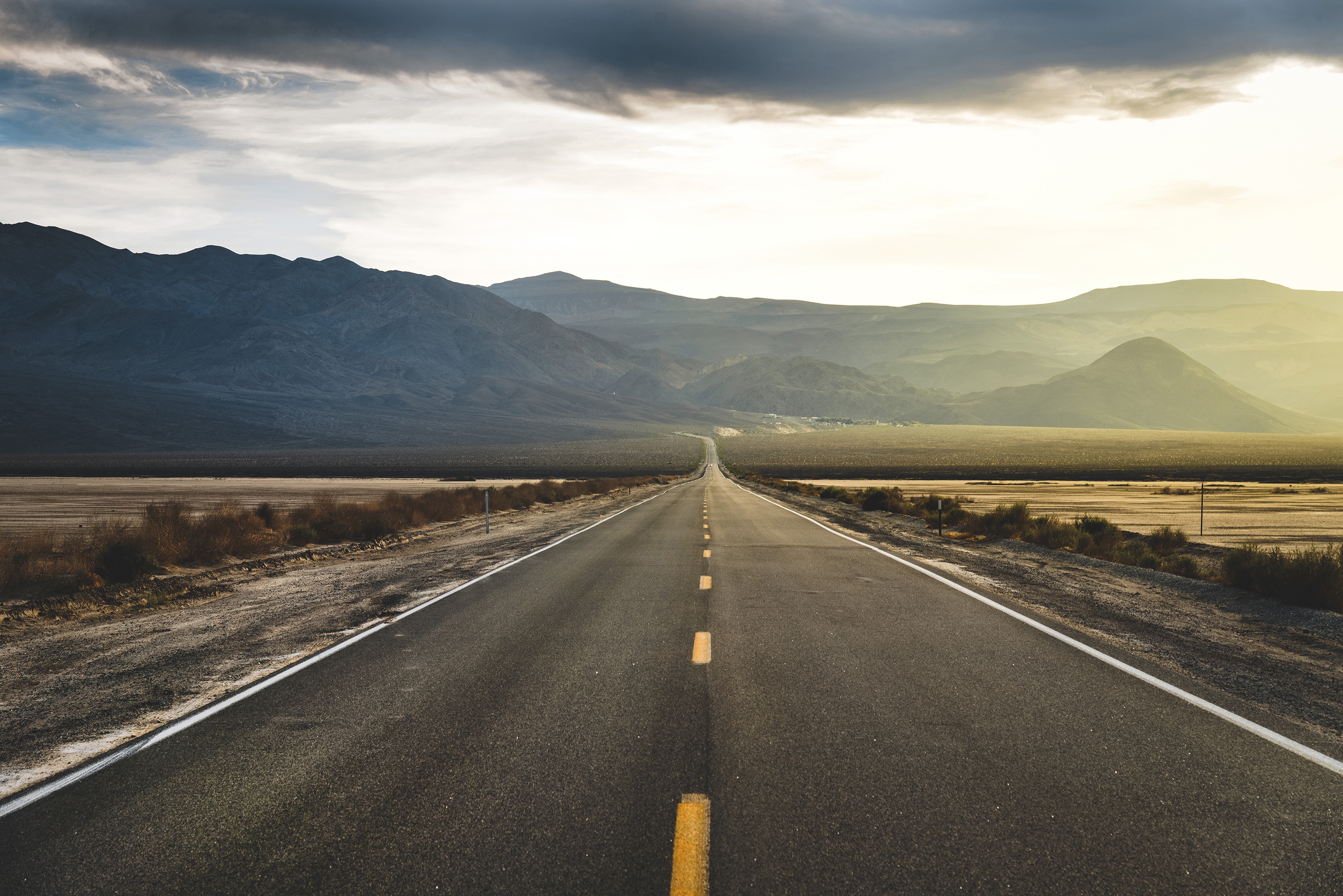 A long road through flat plains with a row of mountains and the rising sun on the horizon