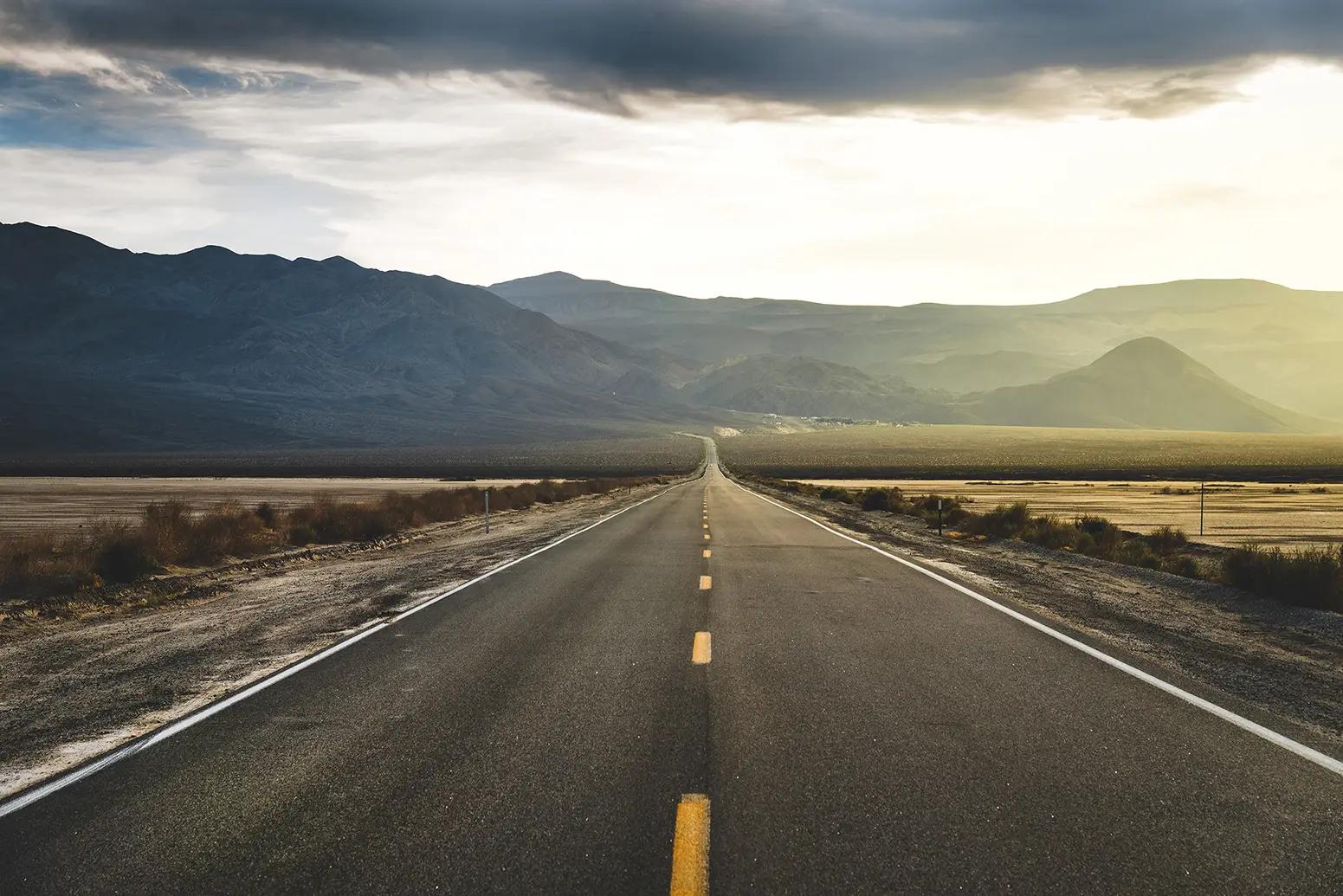 A long road through flat plains with a row of mountains and the rising sun on the horizon
