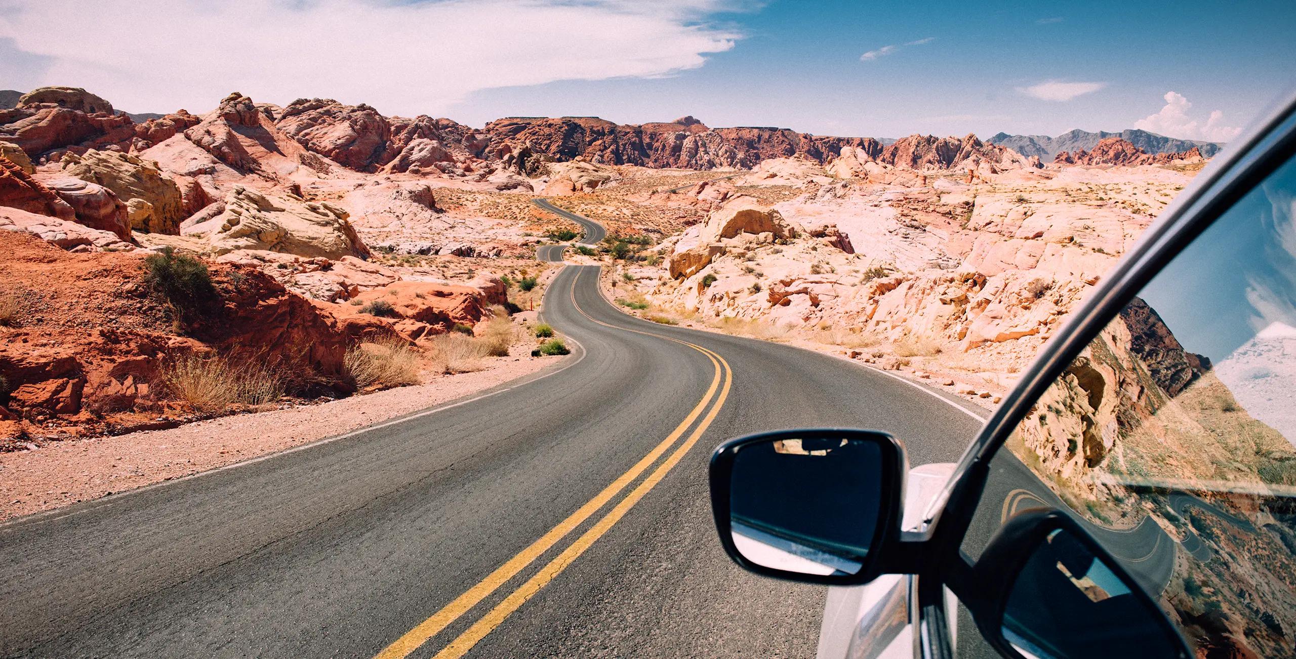 View from a car window of a winding two-lane desert highway cutting through red sandstone rock formations under a partly cloudy blue sky, with the vehicle's side mirror visible in the foreground