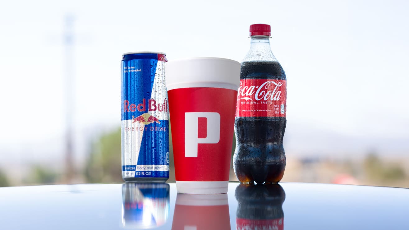 A Red Bull Energy Drink can (12 fl oz), a red and white cup with a Pilot 'P' logo, and a Coca-Cola Original Taste bottle (20 fl oz) displayed on a reflective white surface