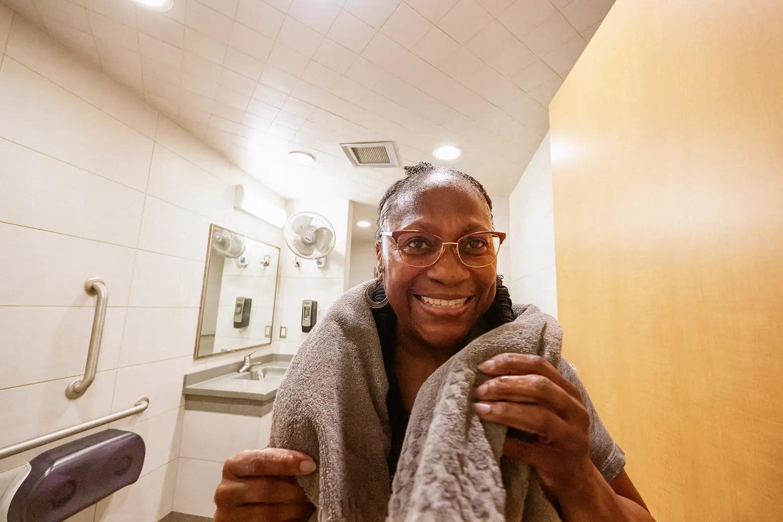 A smiling woman wearing glasses wraps herself in a gray towel inside a clean, well-lit shower facility restroom with a sink and mirror visible in the background