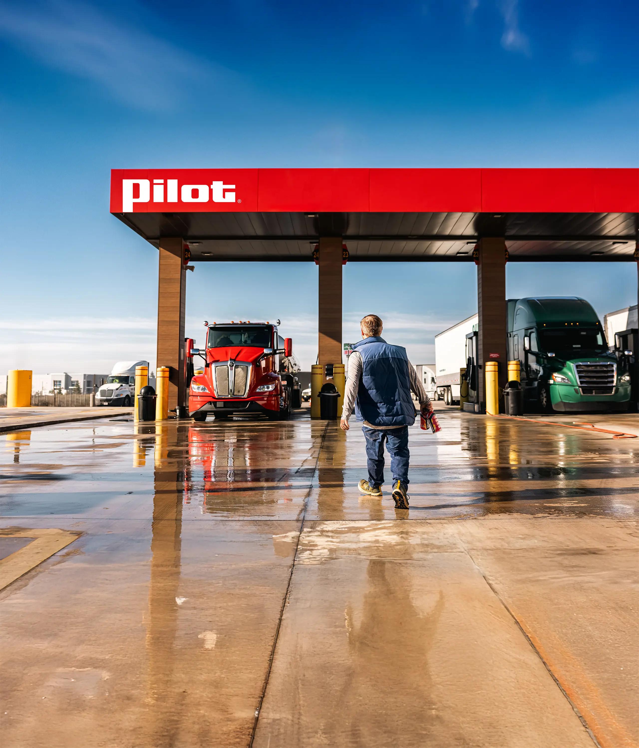 Pro driver walking toward a red semi-truck at a Pilot Travel Center fuel pump on a wet concrete lot with other trucks visible in the background