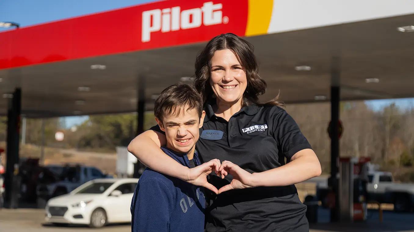 A Pilot Company employee named Hannah and a young boy make a heart shape together with their hands while smiling in front of a Pilot gas station canopy