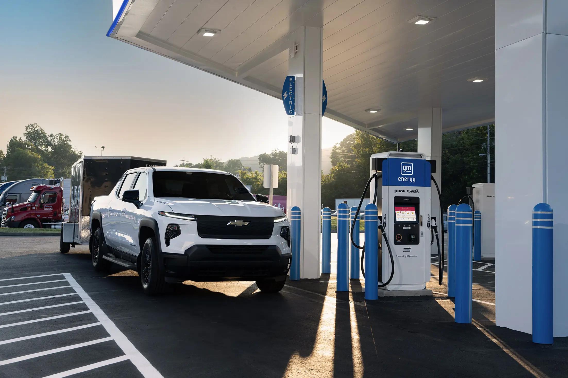 A white EV pickup truck towing a trailer pulls into a covered GM Energy fast-charging station at a Pilot Flying J, with blue DC fast-charger pedestals visible under the canopy