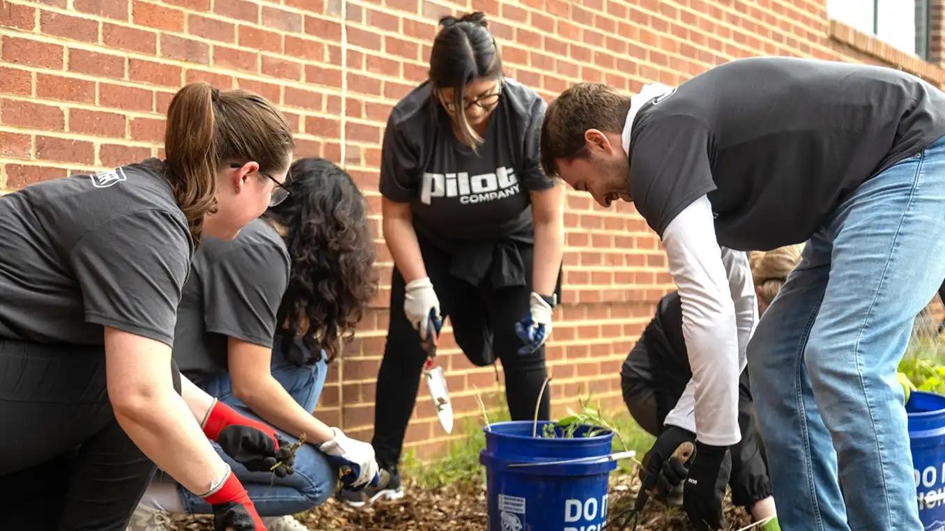 A group of Pilot Company team members wearing branded shirts and gardening gloves planting or weeding a garden bed along a brick building exterior, with blue buckets and hand trowels visible