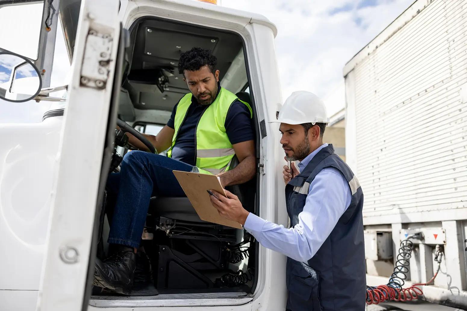 A truck driver in a yellow safety vest sitting in a truck cab reviews a clipboard held by a supervisor wearing a white hard hat and navy work vest, at a loading dock or freight yard.