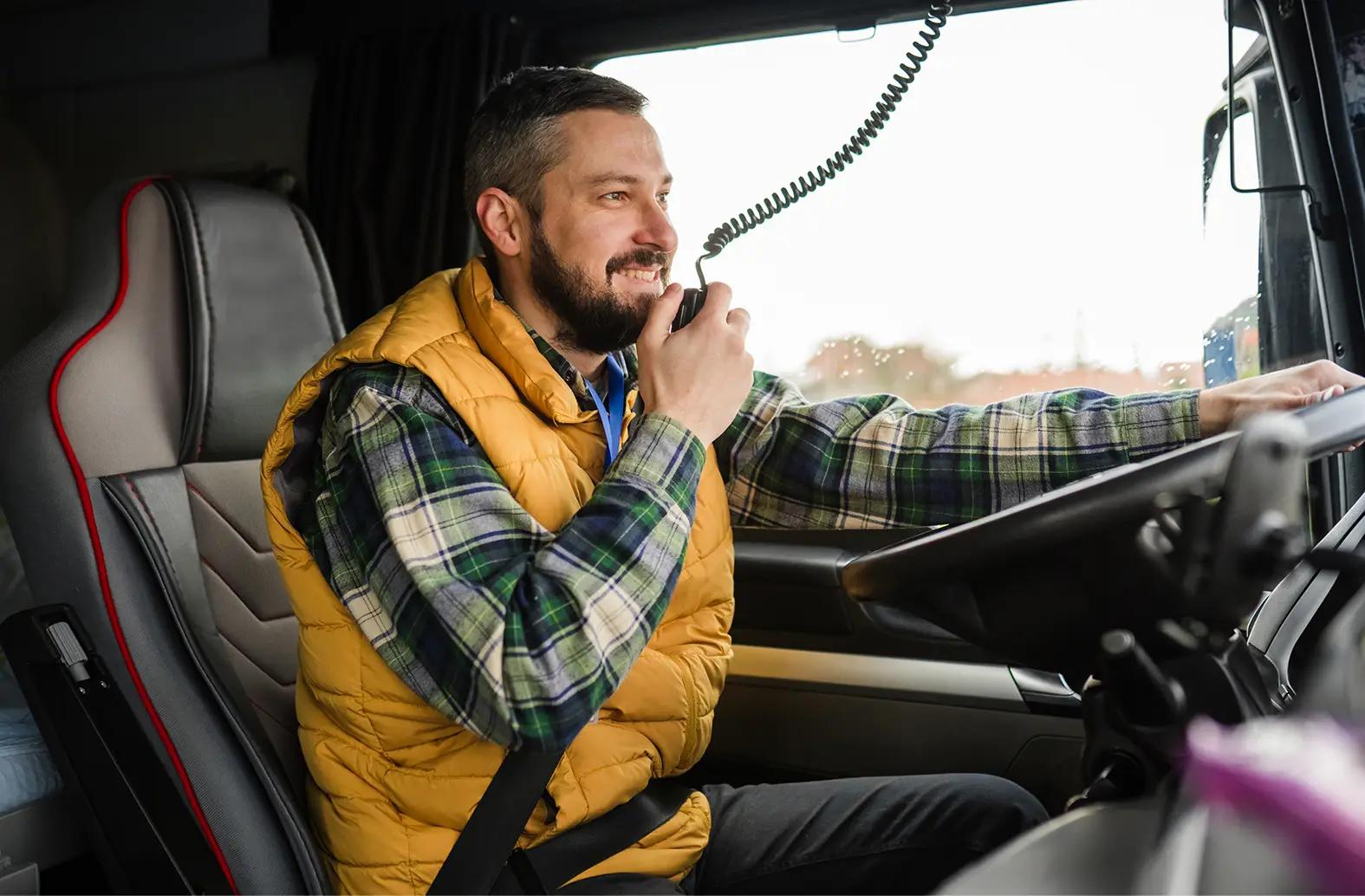 Smiling male truck driver wearing a yellow vest and plaid shirt, sitting in a truck cab with seatbelt fastened, speaking into a CB radio handset while resting one hand on the steering wheel