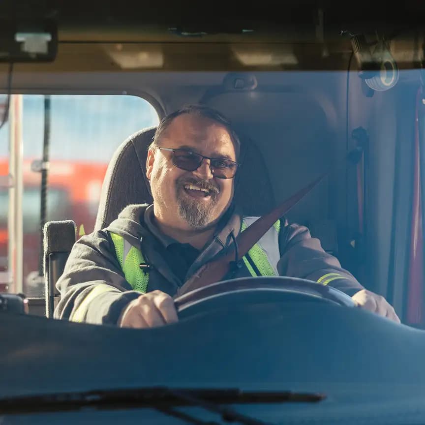 Smiling truck driver wearing a high-visibility safety vest and glasses, seated behind the steering wheel in a truck cab with seatbelt fastened, photographed through the windshield