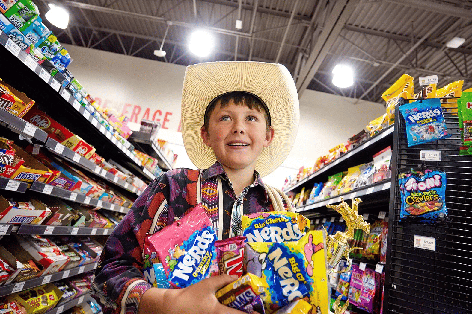 A smiling boy in a cowboy hat and western shirt holds an armful of Nerds candy bags and a gold trophy while standing in the candy aisle of a Pilot store