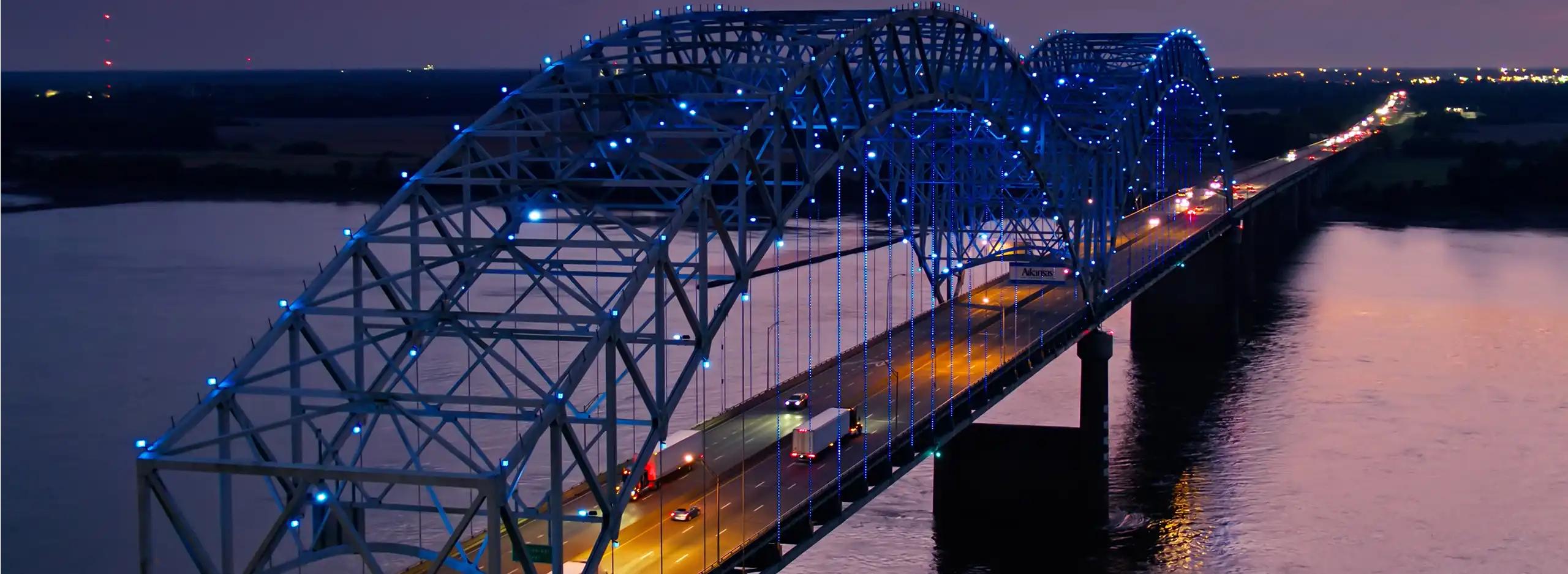 Semi trucks driving over an illuminated bridge at dusk over purple water