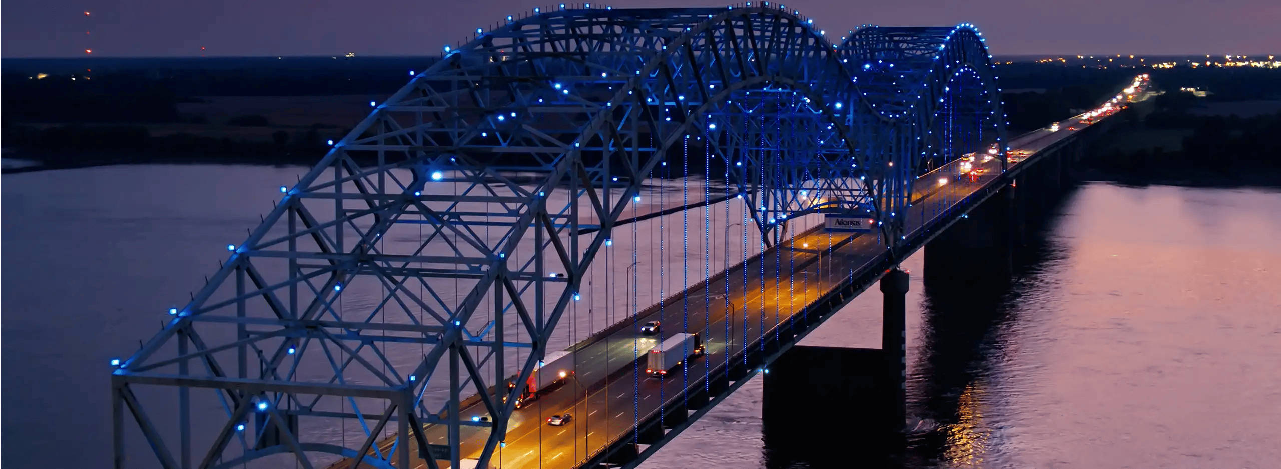 A steel arch bridge illuminated with blue LED lights at dusk, carrying light traffic including one semi truck, spanning a wide dark river with city lights visible on the distant shoreline