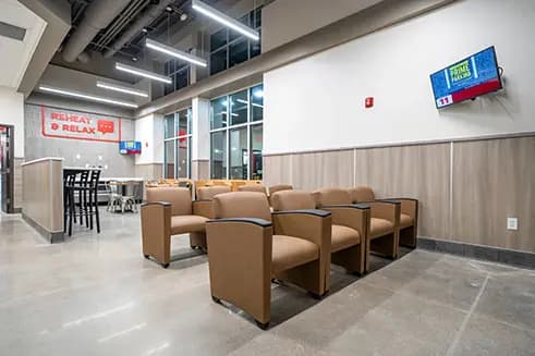 A driver lounge area with two rows of tan upholstered armchairs, a wall-mounted TV, high-top tables with black chairs in the background, and a 'Reheat & Relax' sign on the wall