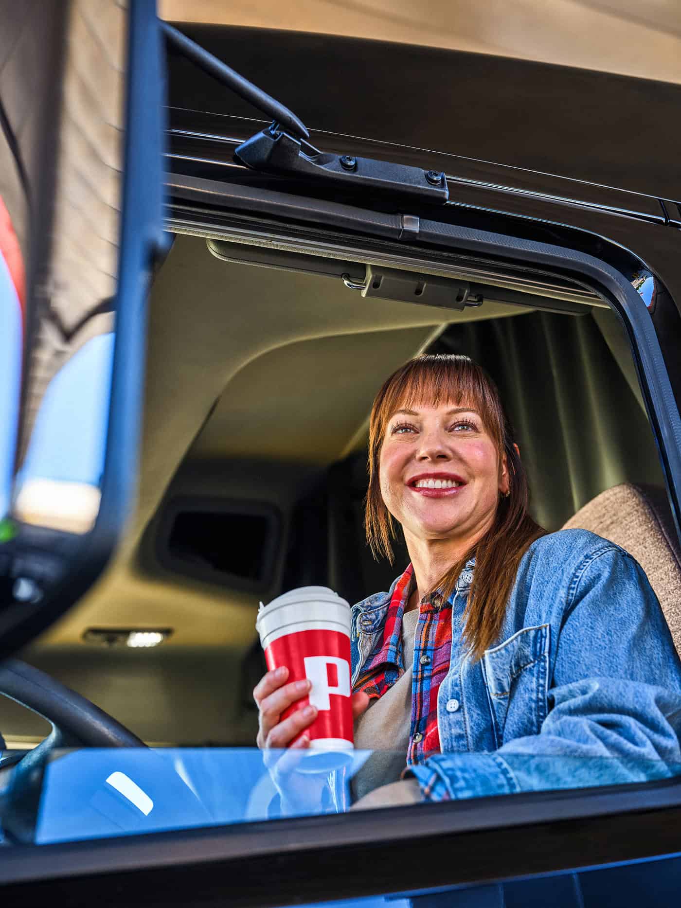 A smiling middle-aged woman with auburn hair sitting in a truck cab, holding a red Pilot branded travel cup with a white lid, wearing a denim jacket over a plaid shirt