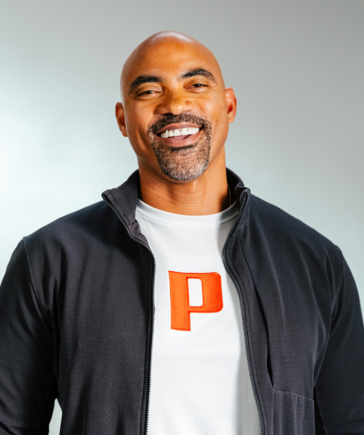 Adam Wright smiling, wearing a white t-shirt with an red Pilot "P" logo and a dark zip-up jacket, professional headshot