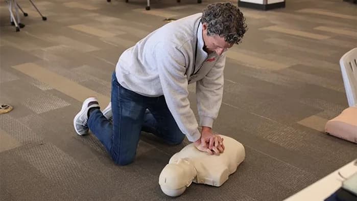 A person kneeling on the floor performing chest compressions on a CPR training mannequin during a first aid training session