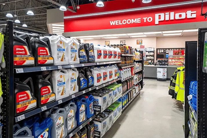 Shelves stocked with motor oil and automotive fluids including Shell Rotella and Pilot Flying J branded oils in various viscosity grades, inside a Pilot travel center store with a 'Welcome to Pilot' sign overhead