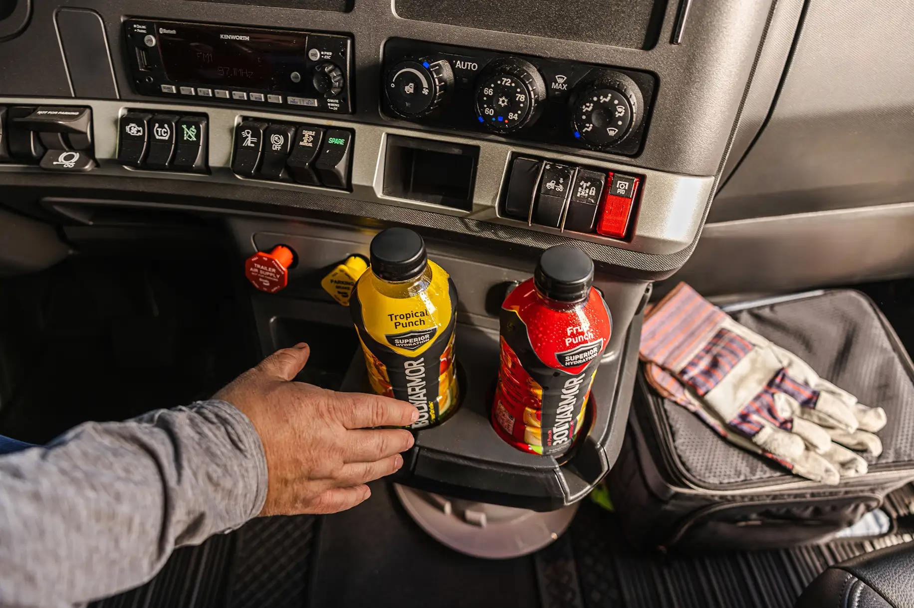 Hand reaching for a BODYARMOR Tropical Punch sports drink in the cup holder of a Kenworth semi truck cab, alongside a BODYARMOR Fruit Punch bottle