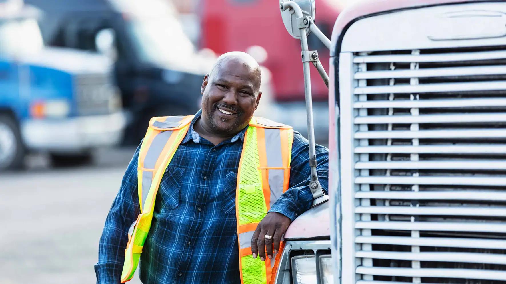 Smiling male truck driver wearing a high-visibility safety vest leaning against the front grille of a semi truck in a commercial trucking yard