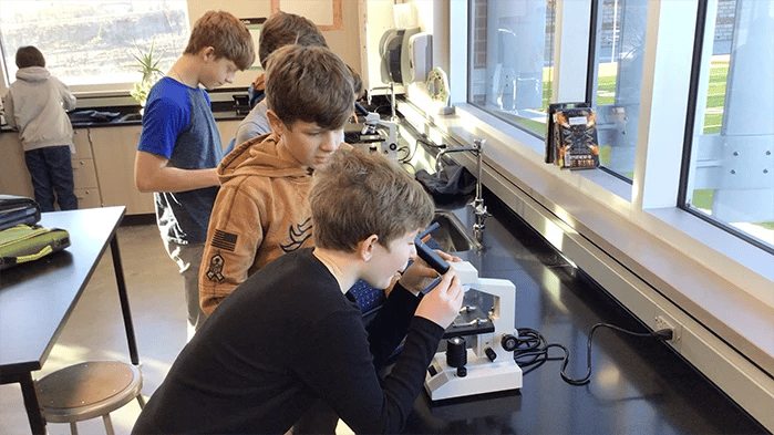 Middle school students using microscopes during a science lab class, with one student leaning in to look through a microscope while classmates observe nearby