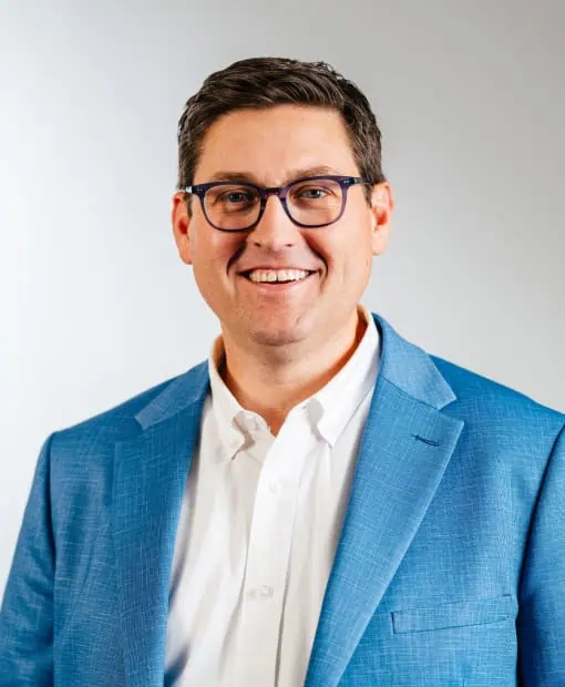 Professional headshot of Josh Birdwell, a man wearing a blue blazer over a white button-down shirt and dark-framed glasses, smiling against a light gray background.