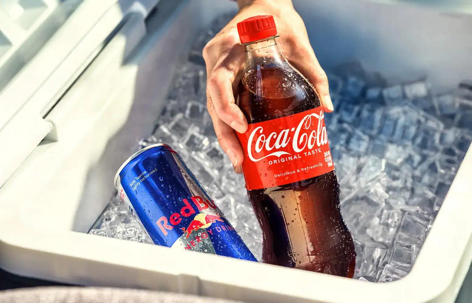 Hand reaching into cooler to grab a Coca-Cola with a Red Bull can in the ice chest.