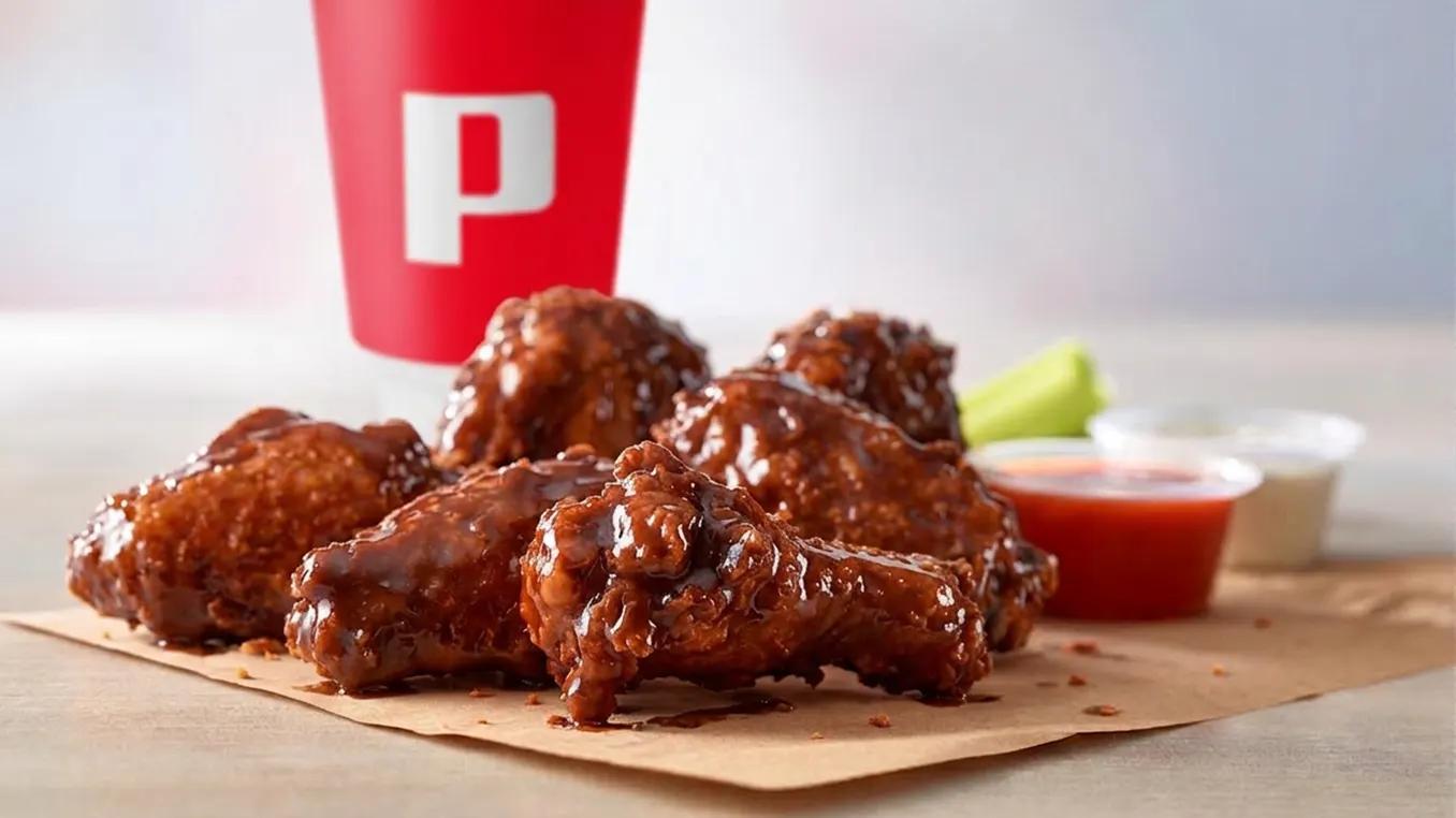 Saucy BBQ glazed chicken wings on parchment paper, served with dipping sauces and celery, with a red Popeyes branded cup in the background