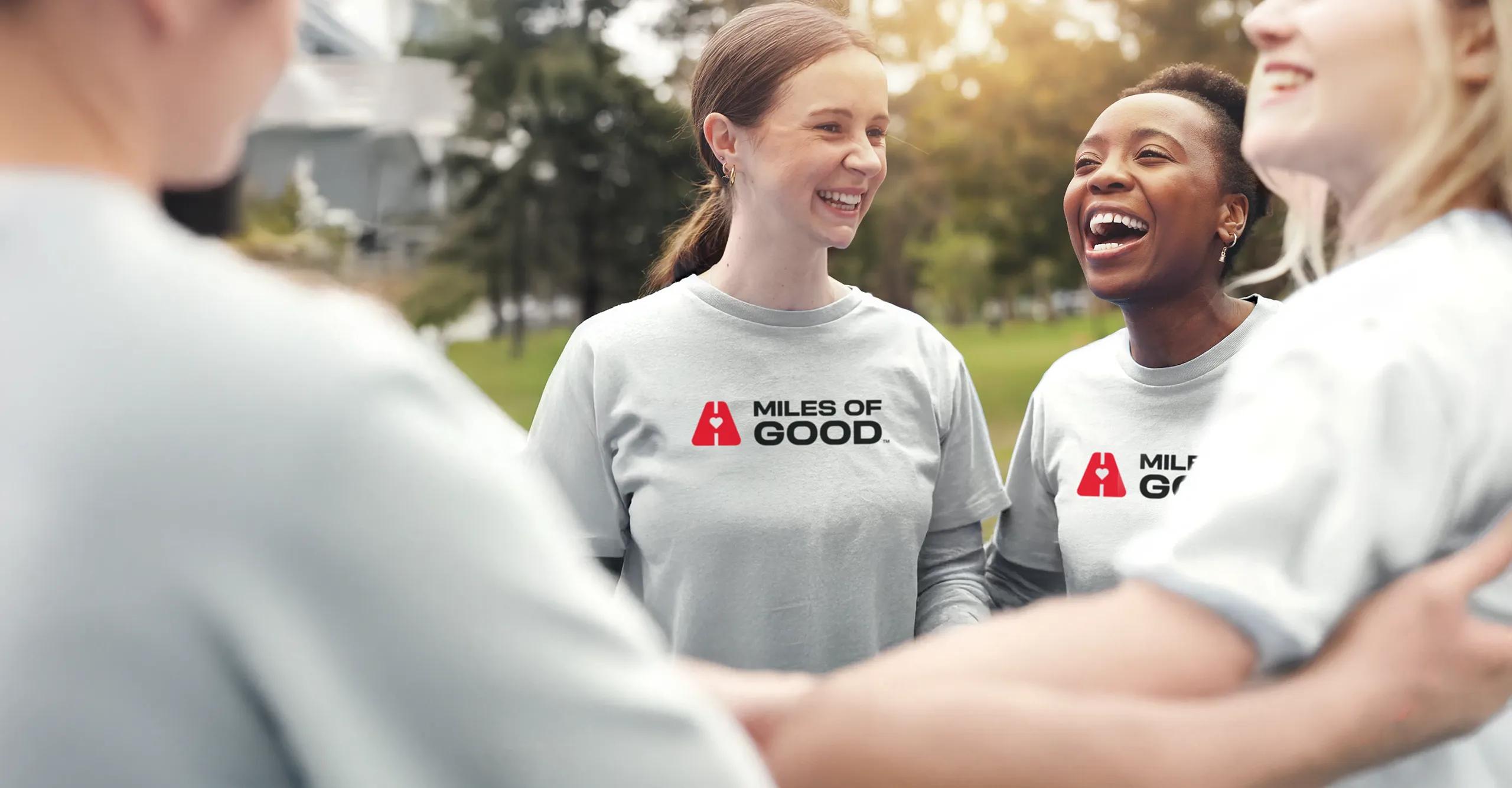 A group of smiling and laughing volunteers in a team huddle outdoors, wearing grey Miles of Good‚ long-sleeve shirts in a park setting