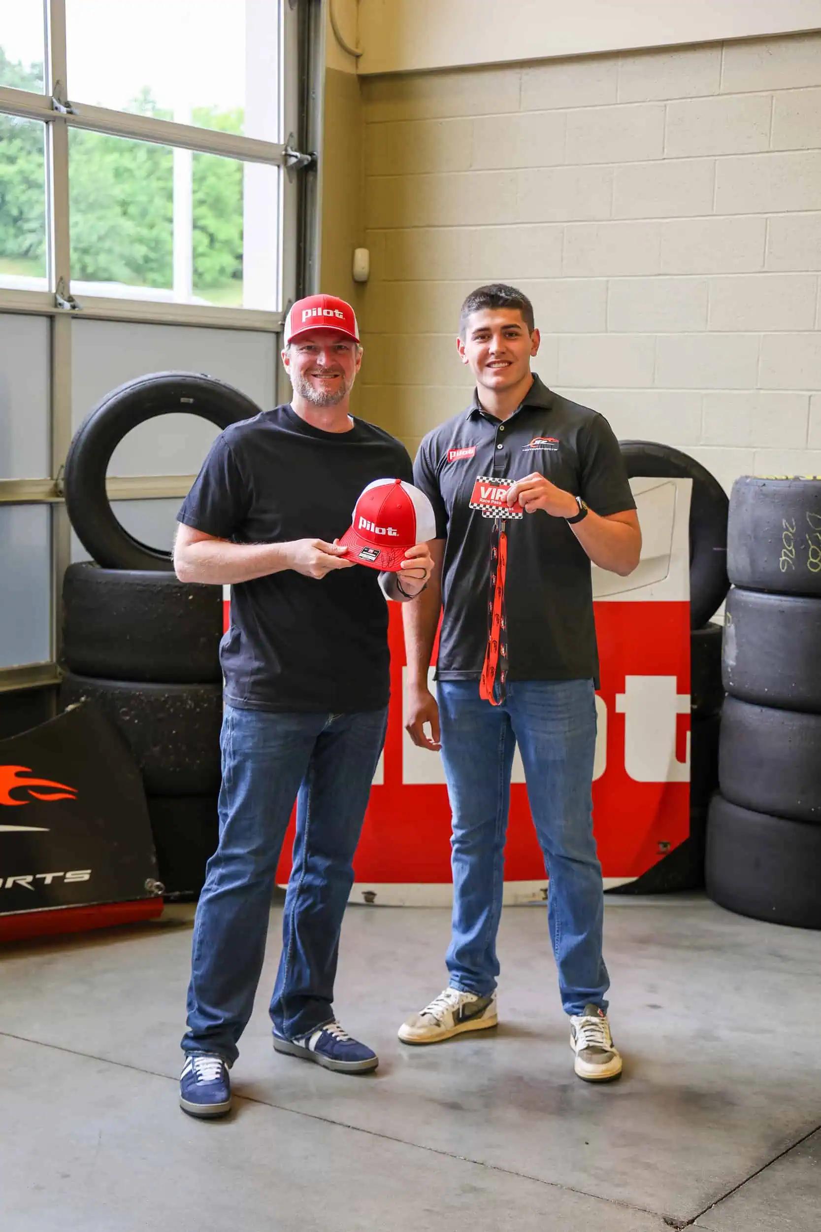Dale Earnhardt Jr. and Sammy Smith stand in a racing garage holding Pilot-branded items, including a red-and-white Pilot hat and a VIP pass. Stacks of racing tires and Pilot signage are visible in the background.