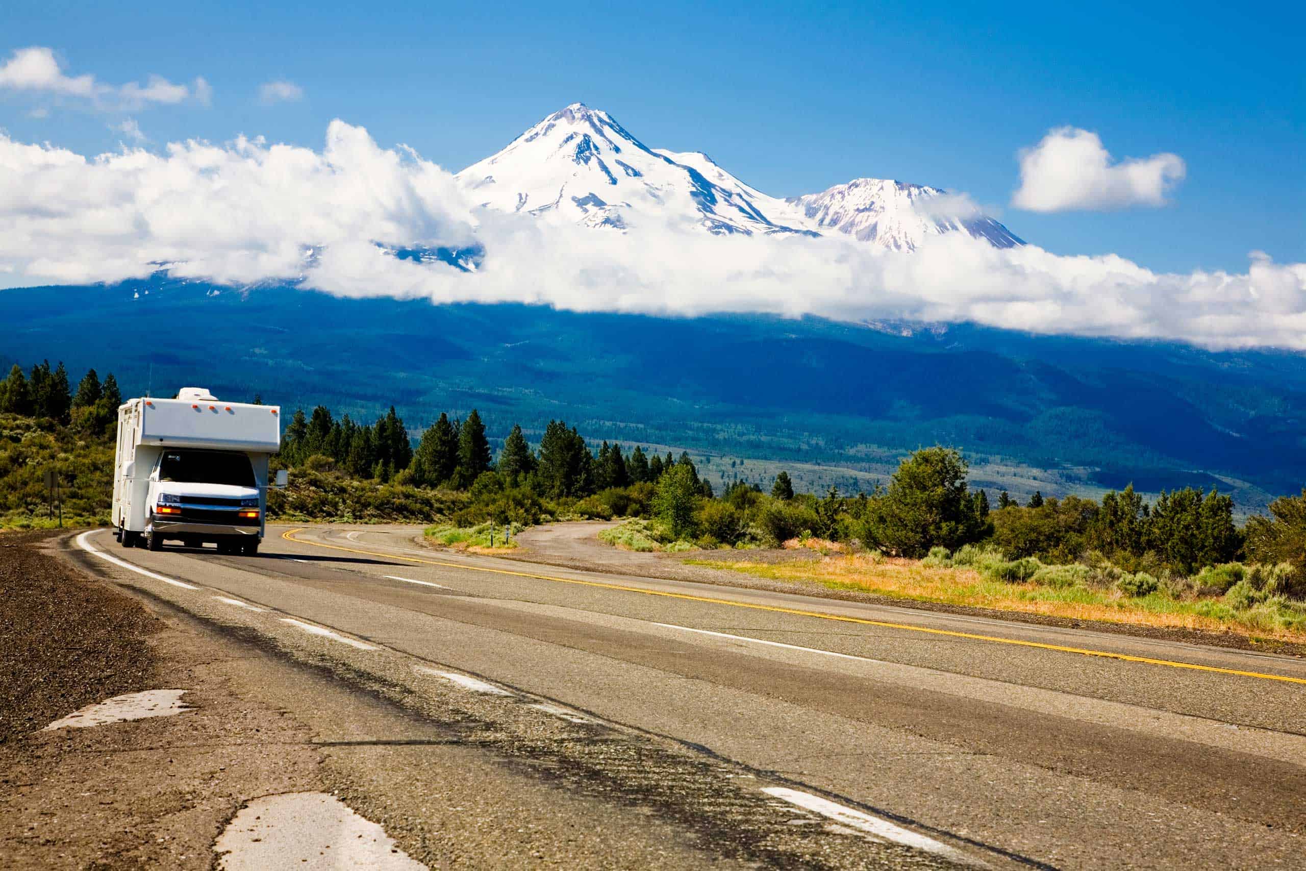 A white class C motorhome traveling along a winding two-lane highway through a forested landscape, with snow-capped Mount Shasta rising above the clouds in the background under a bright blue sky