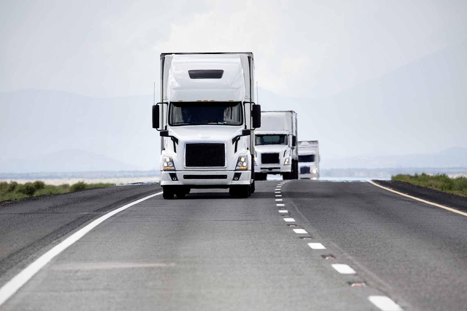 Fleet of white semi trucks driving down a long paved road with a dashed white line down the center