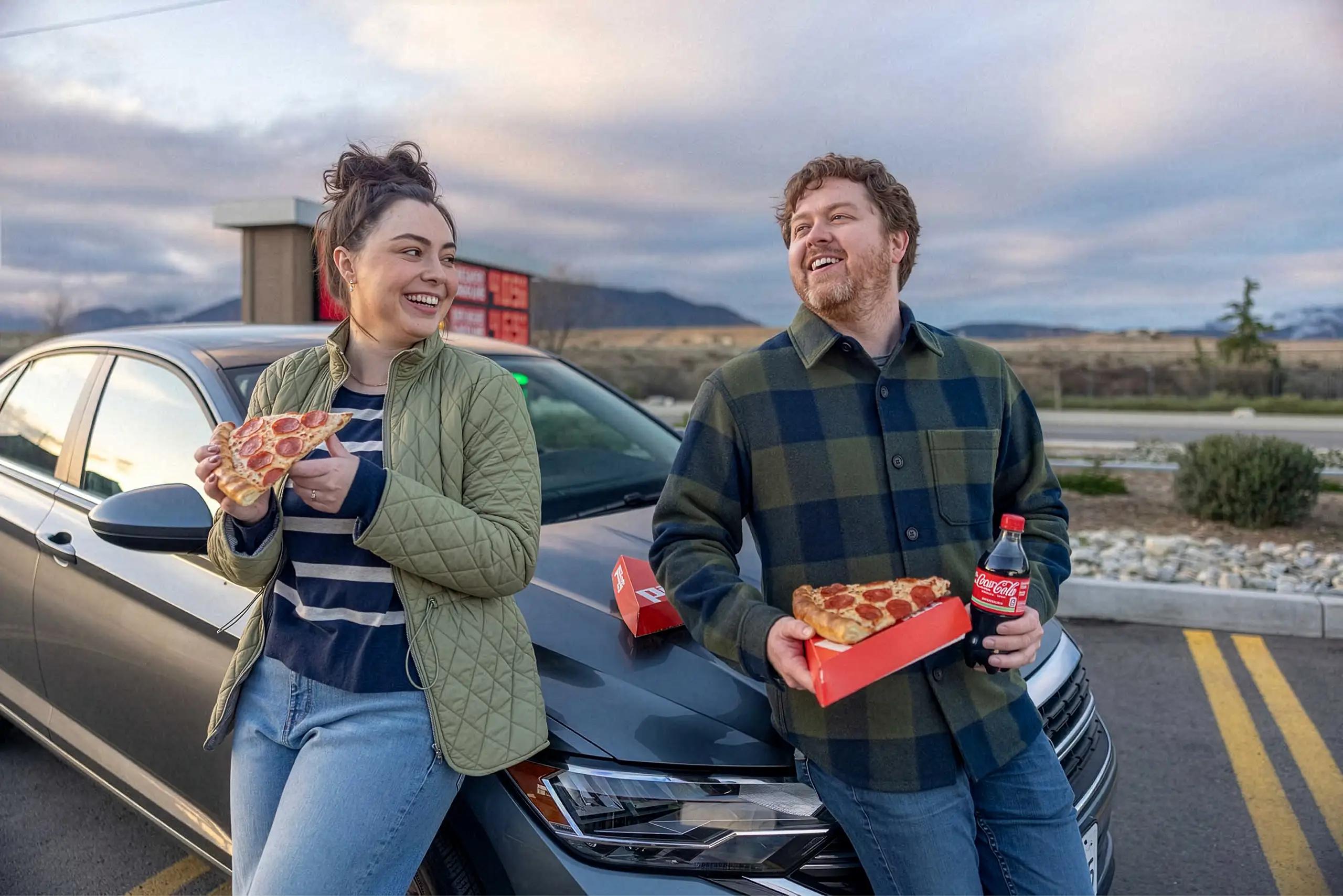 A couple enjoying a slice of Pilot pizza on the hood of their car