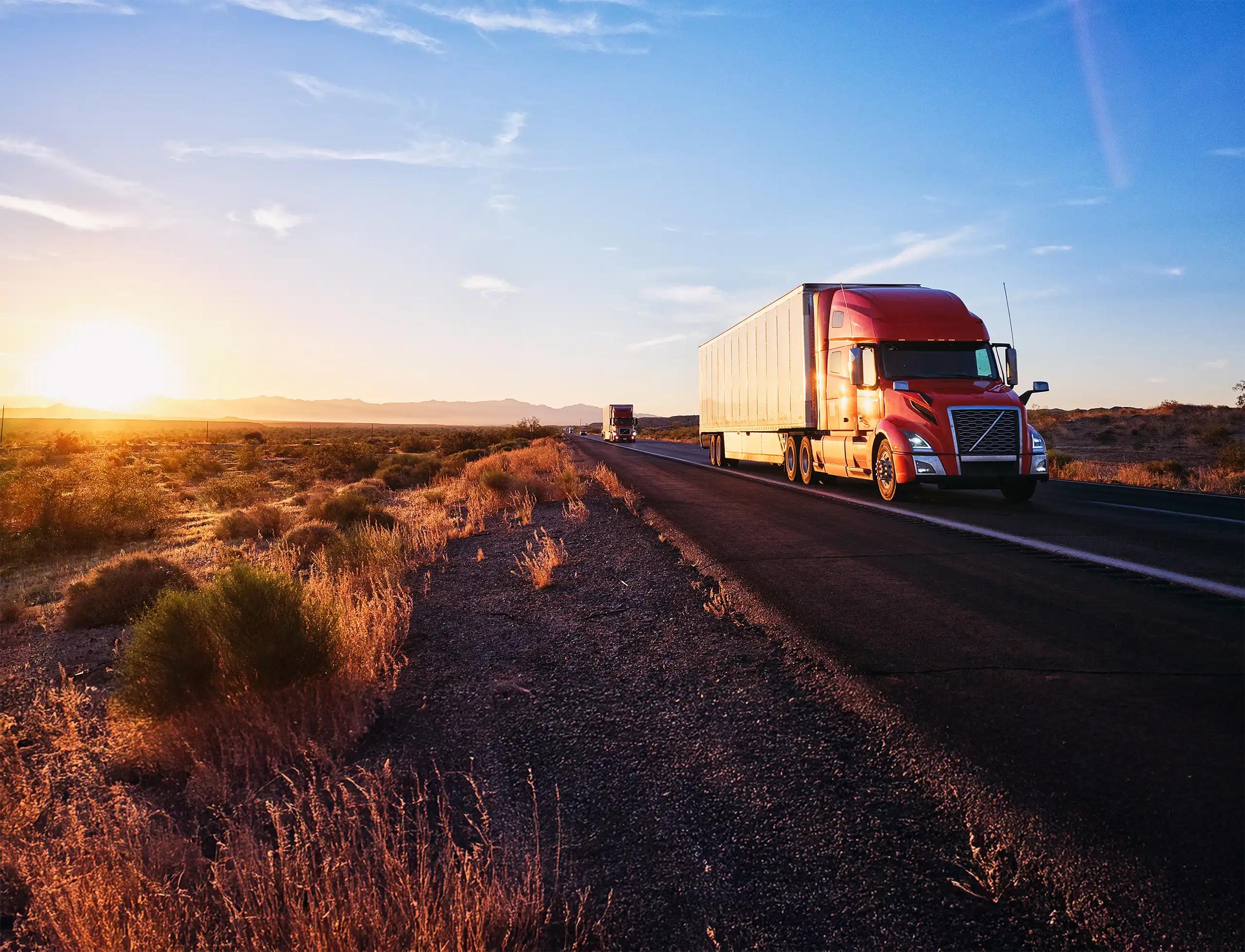 A red semi-truck with a white trailer traveling along a desert highway at sunset, with arid scrubland and mountains visible in the background