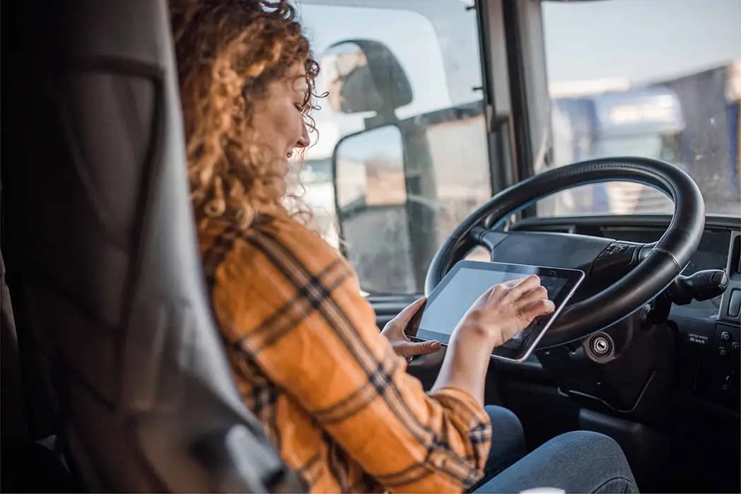 A woman with curly hair wearing a plaid shirt sits in the driver's seat of a large truck cab, smiling while tapping on a tablet device