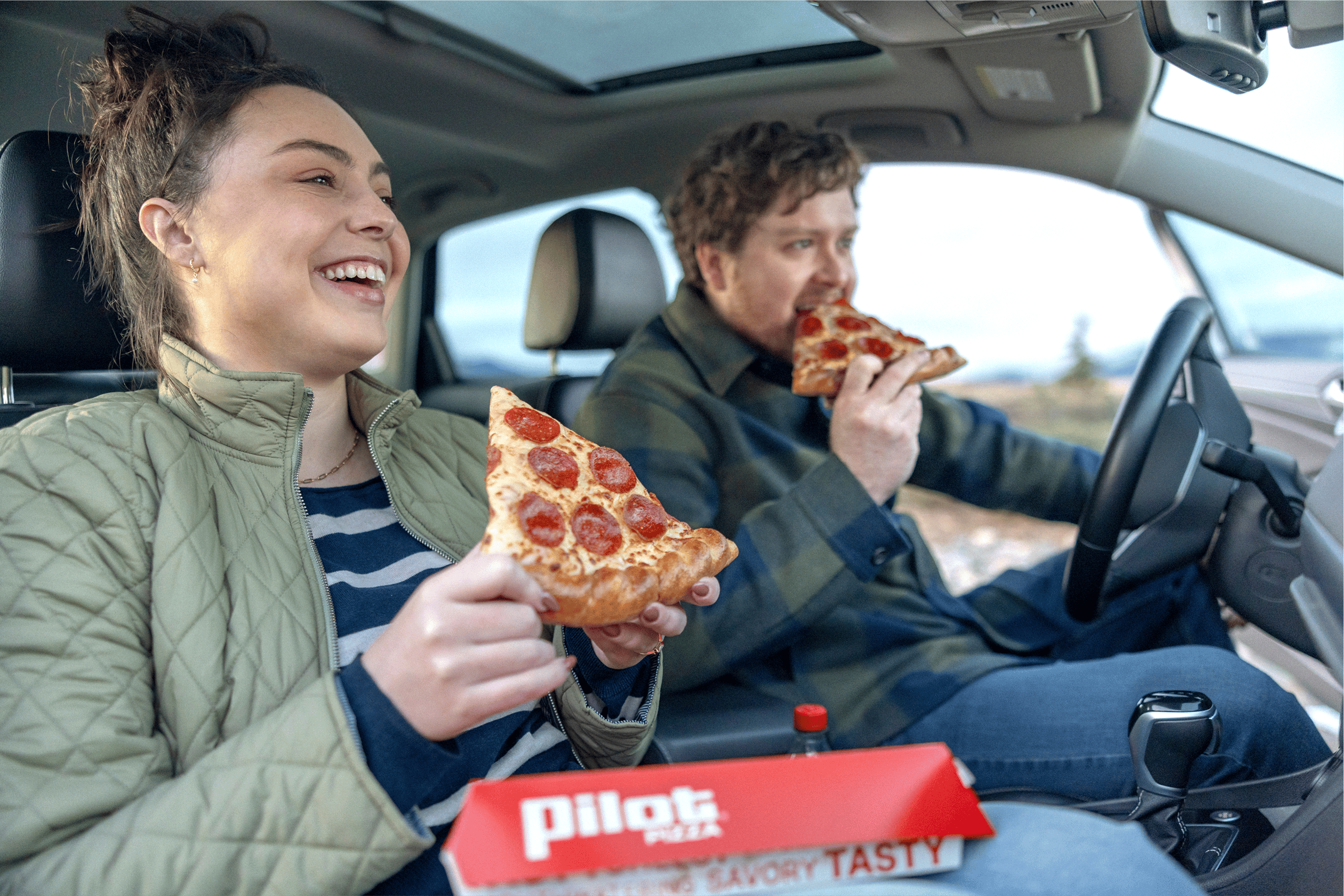 A woman and a man inside a vehicle eating two slices of pepperoni pizza