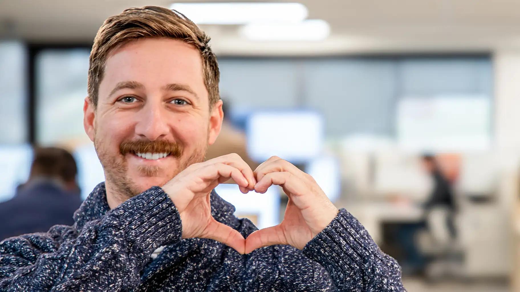 Smiling man in a blue knit sweater making a heart shape with his hands in a bright open office environment