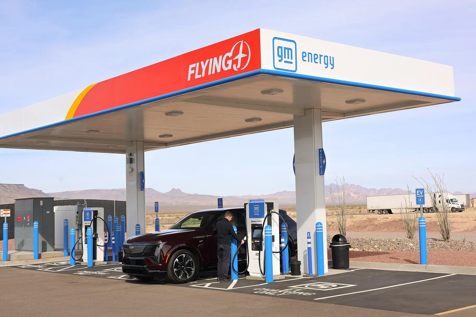 A person charging a maroon electric SUV at a Flying J and GM Energy co-branded EV fast-charging station in a desert highway location, with multiple blue-bollard-protected charging stalls under a canopied structure